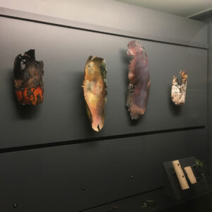 Quatre sculptures murales en métal émaillé de l'artiste Natacha Baluteau, exposées en vitrine. Ces œuvres d'art contemporain en cuivre et émail présentent des textures organiques et des bords irréguliers découpés. De gauche à droite : une pièce ocre et rouge, une beige et verte, une violette et une beige tâchée, évoquant des fragments archéologiques.