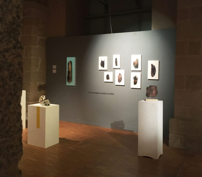 Mise en scène de la série 'Archéo du réel' par Natacha Baluteau à la Galerie des Hospices. L'image montre en premier plan une sculpture murale en émail sur cuivre aux bords déchiquetés, suivie d'une composition de petits formats blancs alignés.