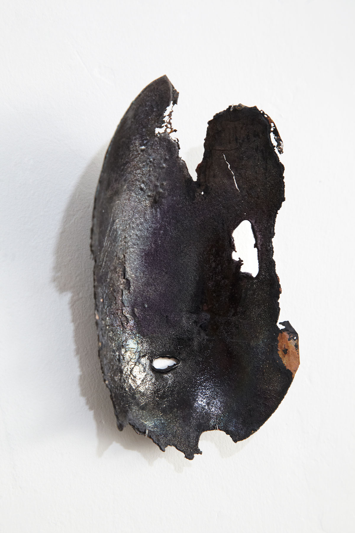 Sculpture murale en émail contemporain sur cuivre de la série Archéo par Natacha Baluteau. Œuvre d’art en forme de carapace sombre et perforée, évoquant un fragment précieux issu de fouilles archéologiques. Lisières de l'objet dentellée proches de l'effritement.