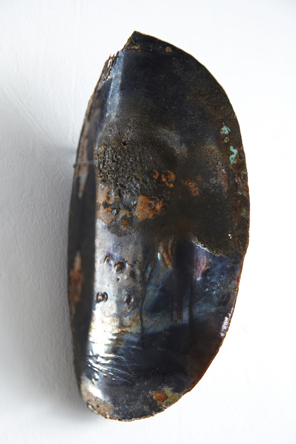 Sculpture murale en émail contemporain sur cuivre, issue de la série Archéo de l'artiste plasticienne Natacha Baluteau. Œuvre d'art unique en forme de coque oblongue aux nuances métalliques sombres et irisations bleutées, témoignant des arts du feu de Limoges.